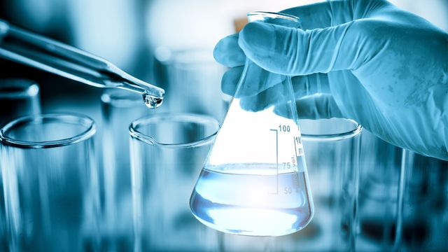 A gloved hand holds a conical flask containing water. A glass pipette containing water can be seen to the left. In the background there are many test tubes.  