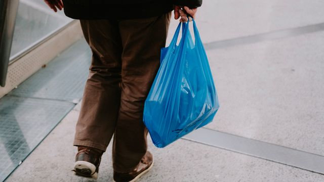 A person in brown trousers carrying a blue plastic bag as they walk down a hallway 