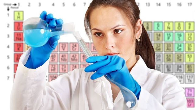 Woman in a lab coat stood in front of the periodic table pouring liquid between two glass flasks. 